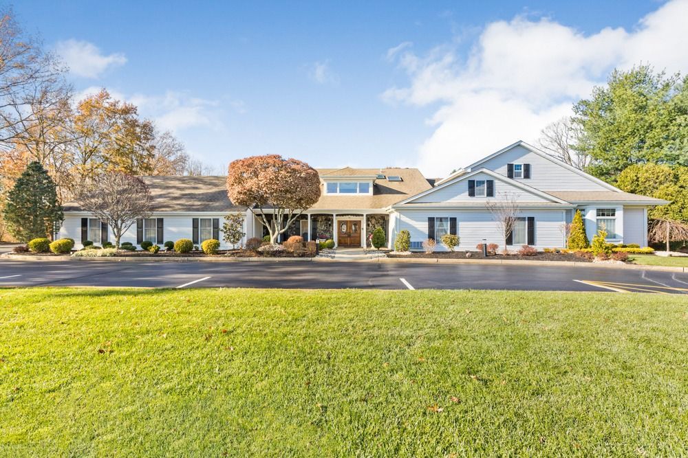 A light-colored, sprawling house with a large parking lot and green lawn under a bright blue sky.