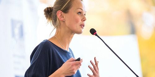 Woman speaking into a microphone during a presentation, gesturing with her hand.