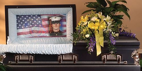 Black casket with American flag and framed military portrait beside yellow flowers