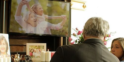 Memorial display with framed photos, candles, and a TV showing an older couple dancing; people stand nearby