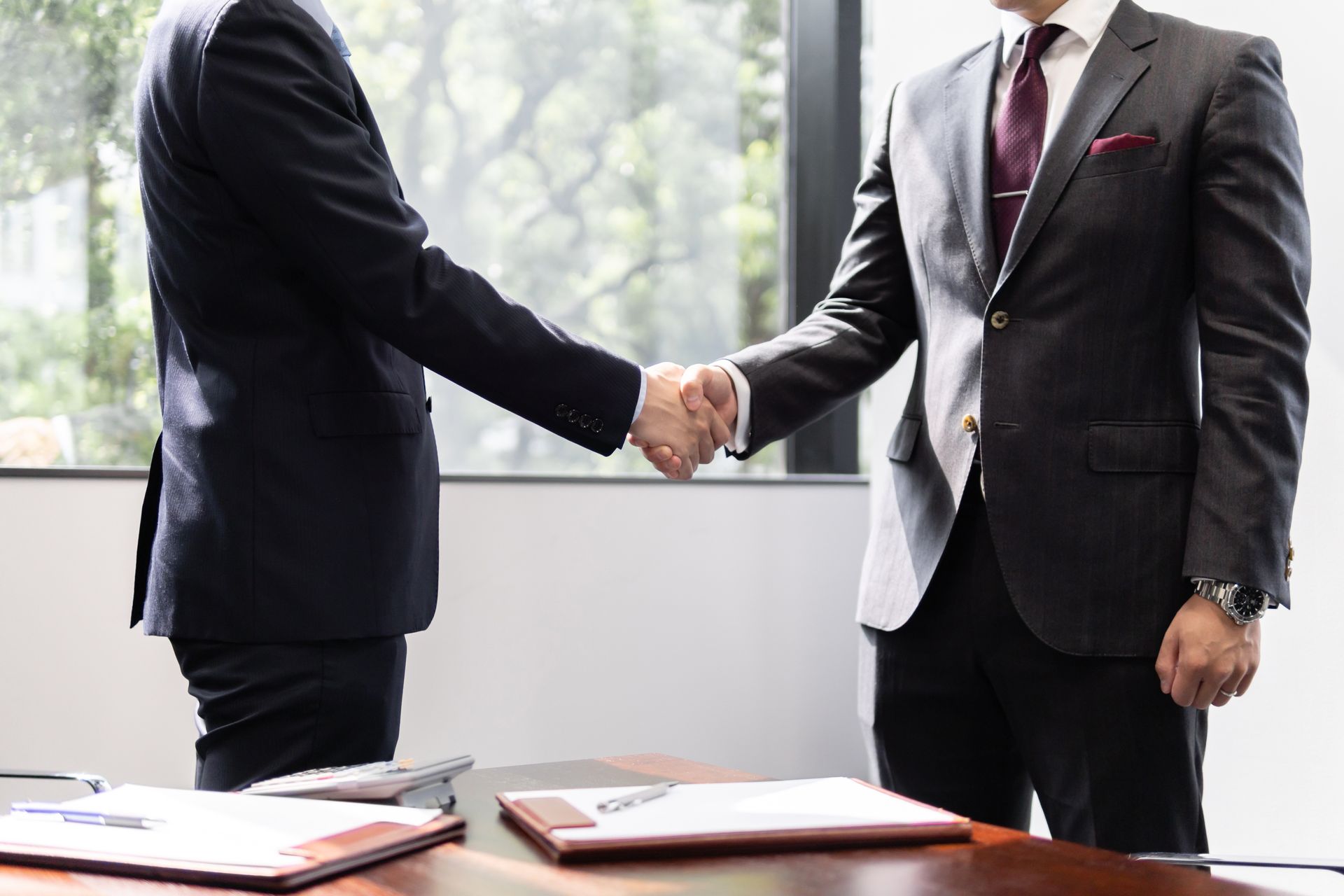 Two businesspeople in dark suits shaking hands across a conference table in a bright office