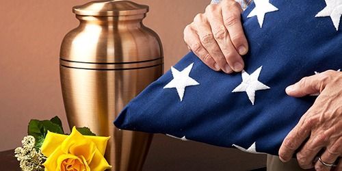 American flag draped over a funeral urn beside yellow flowers and a hand in a solemn memorial scene