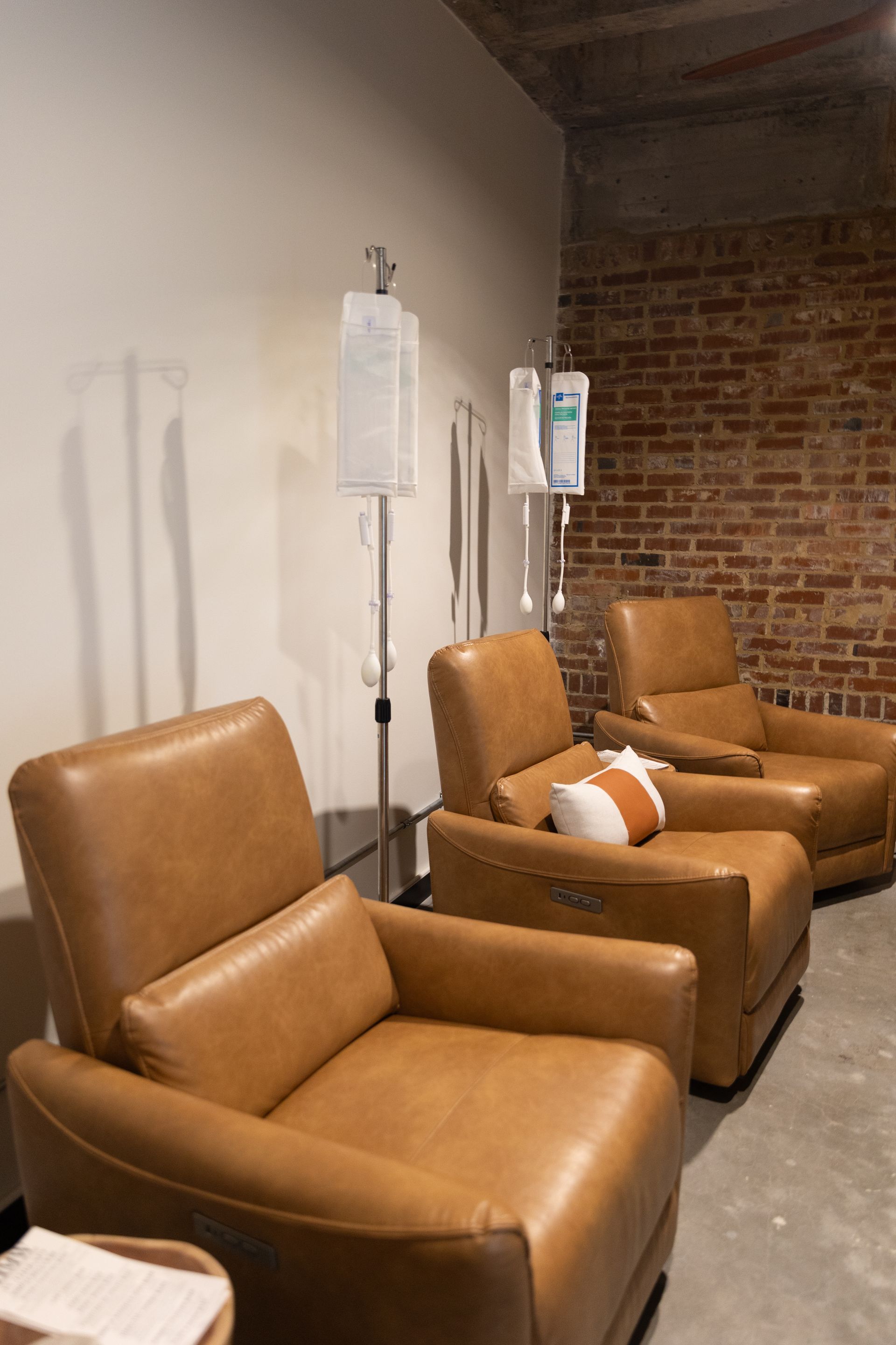 Three brown leather armchairs with IV drips in a room with a brick wall.