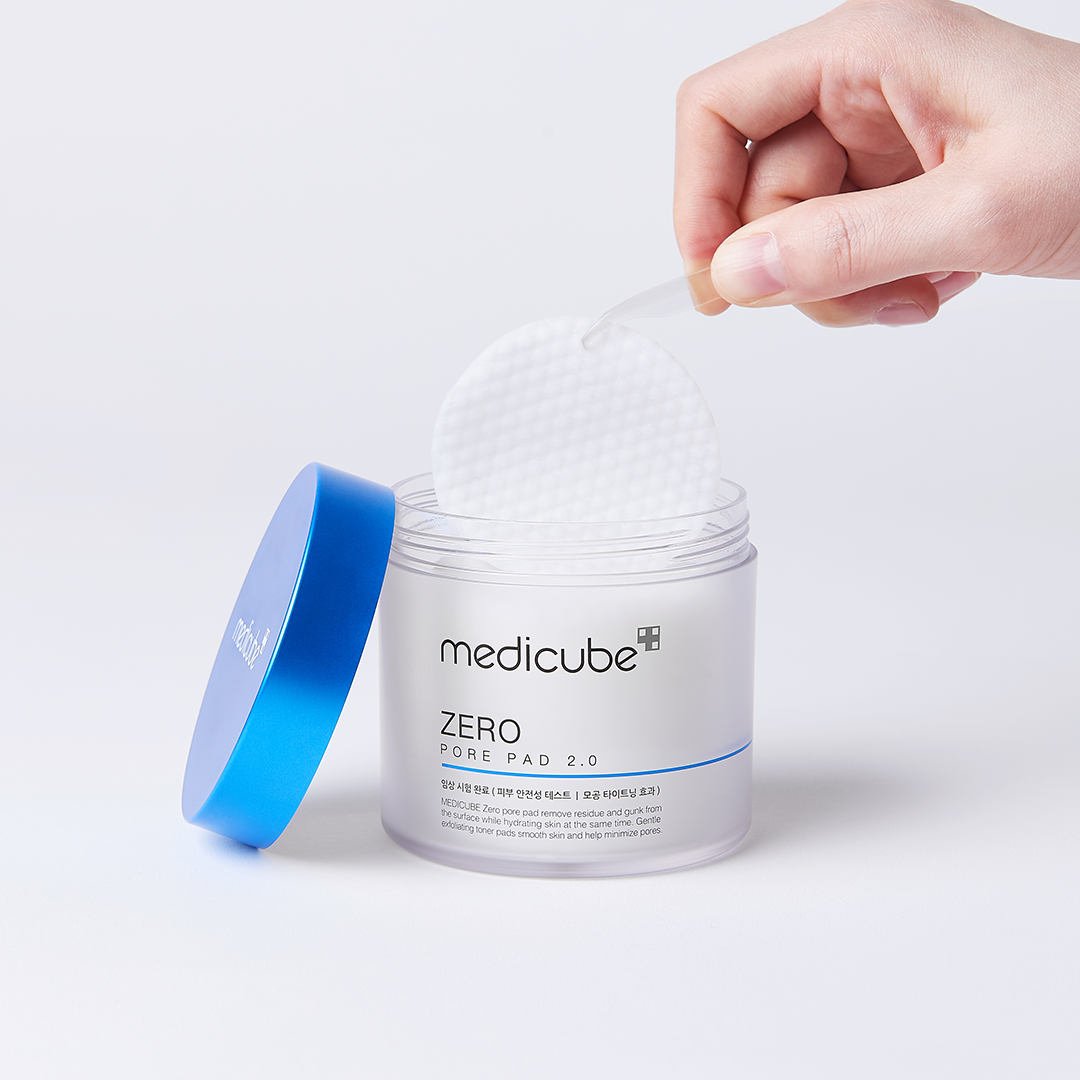 Hand removing a cleansing pad from a medicube Zero pore pad jar with a blue lid on a white background.