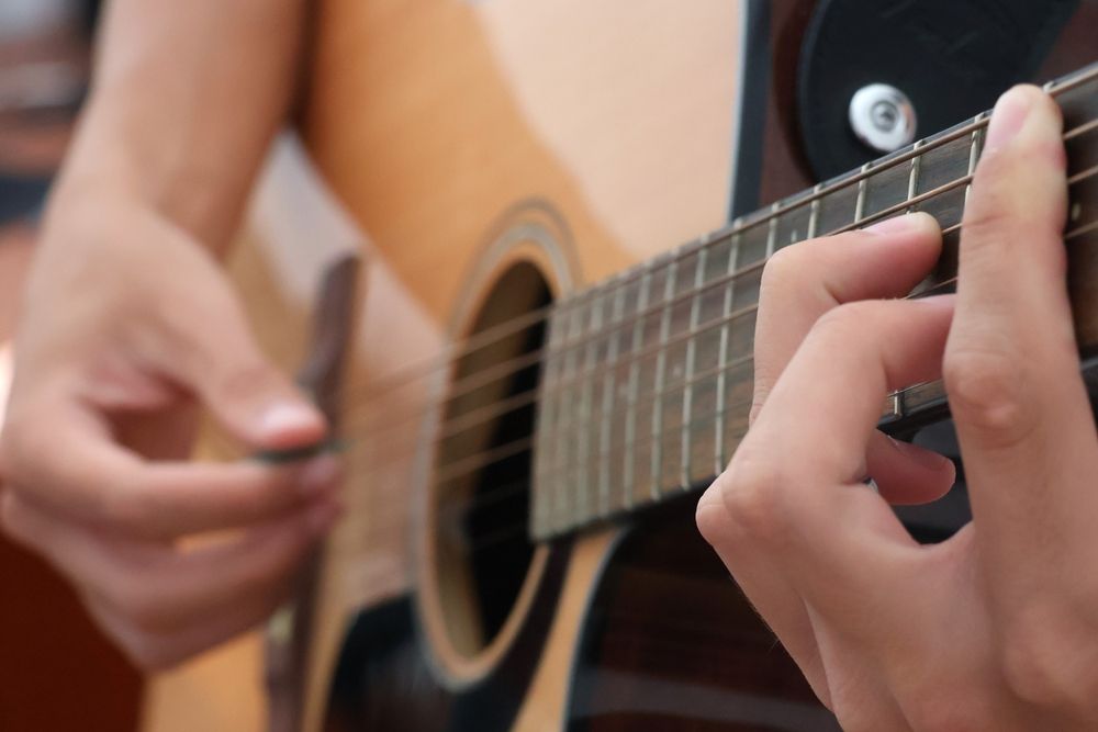 Hands strumming acoustic guitar, fingers on frets, wooden instrument.