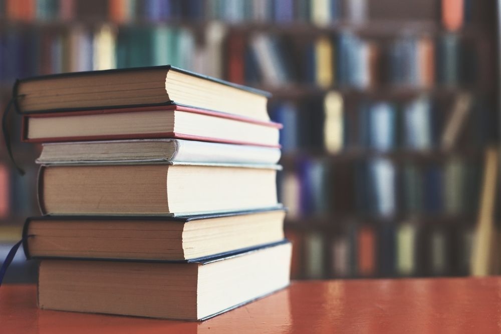 Stack of books on a table with a blurred bookshelf in the background.