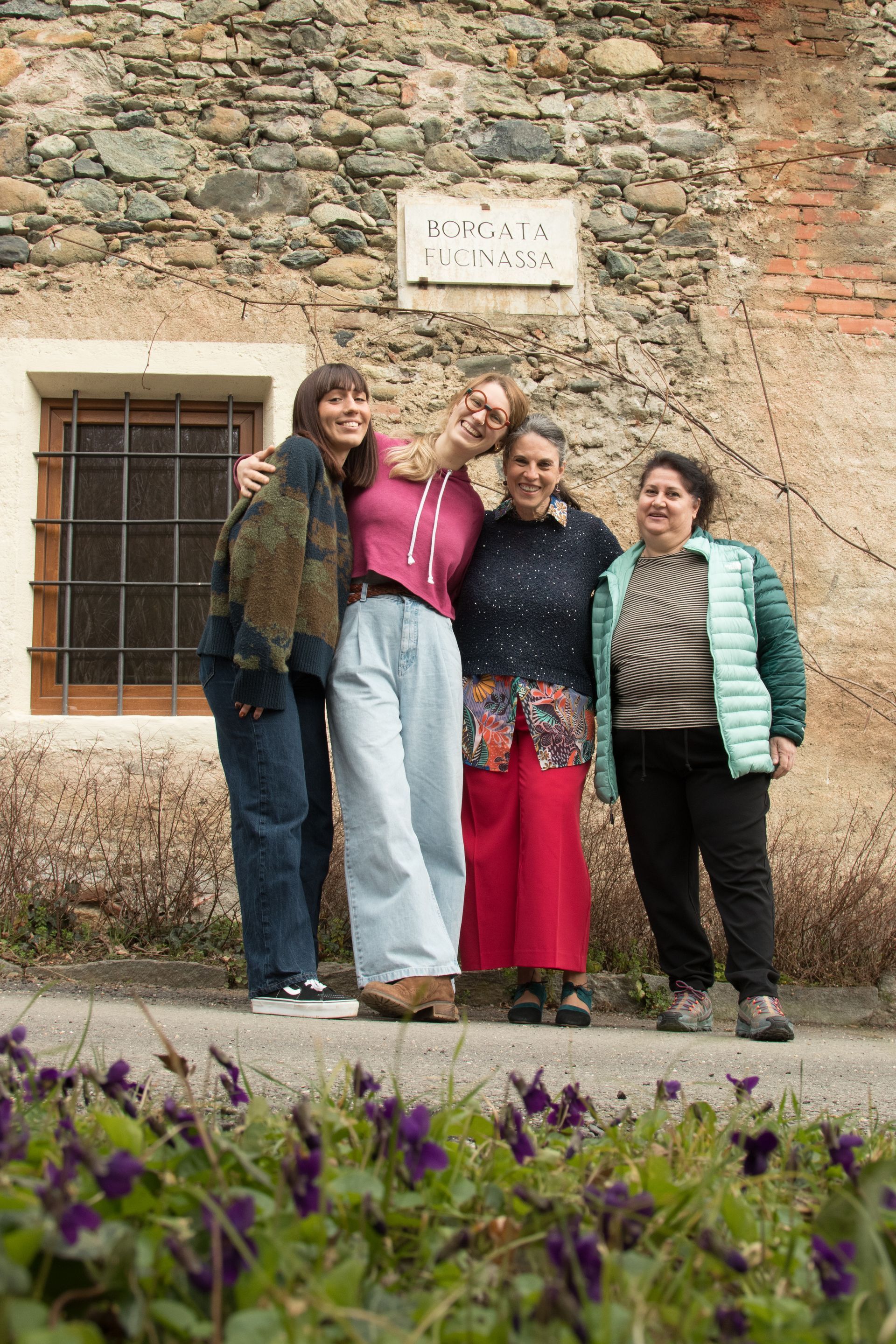 Quattro persone in posa accanto a un muro di pietra con fiori in primo piano.