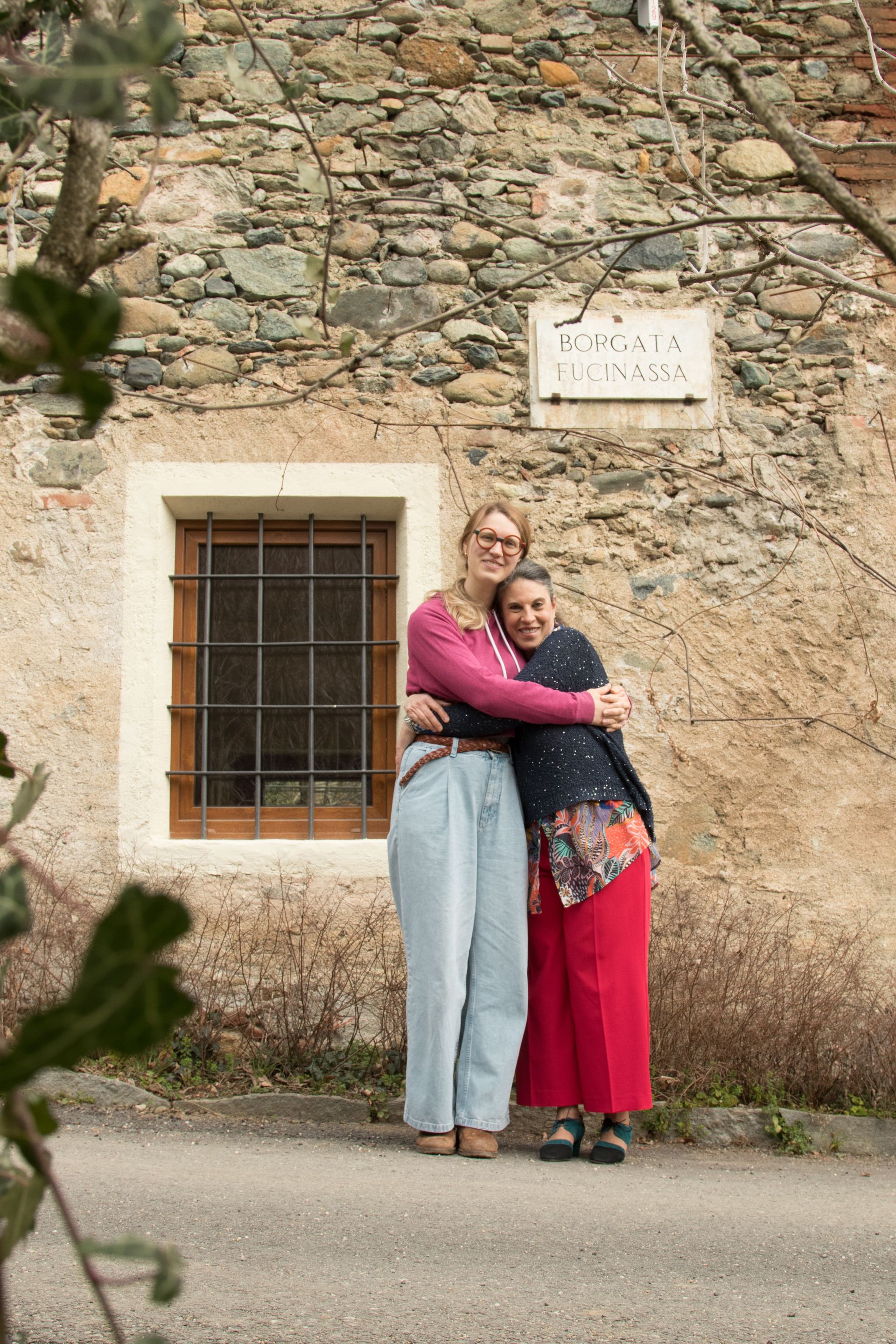 Due persone si abbracciano davanti a un rustico muro di pietra e a una finestra.
