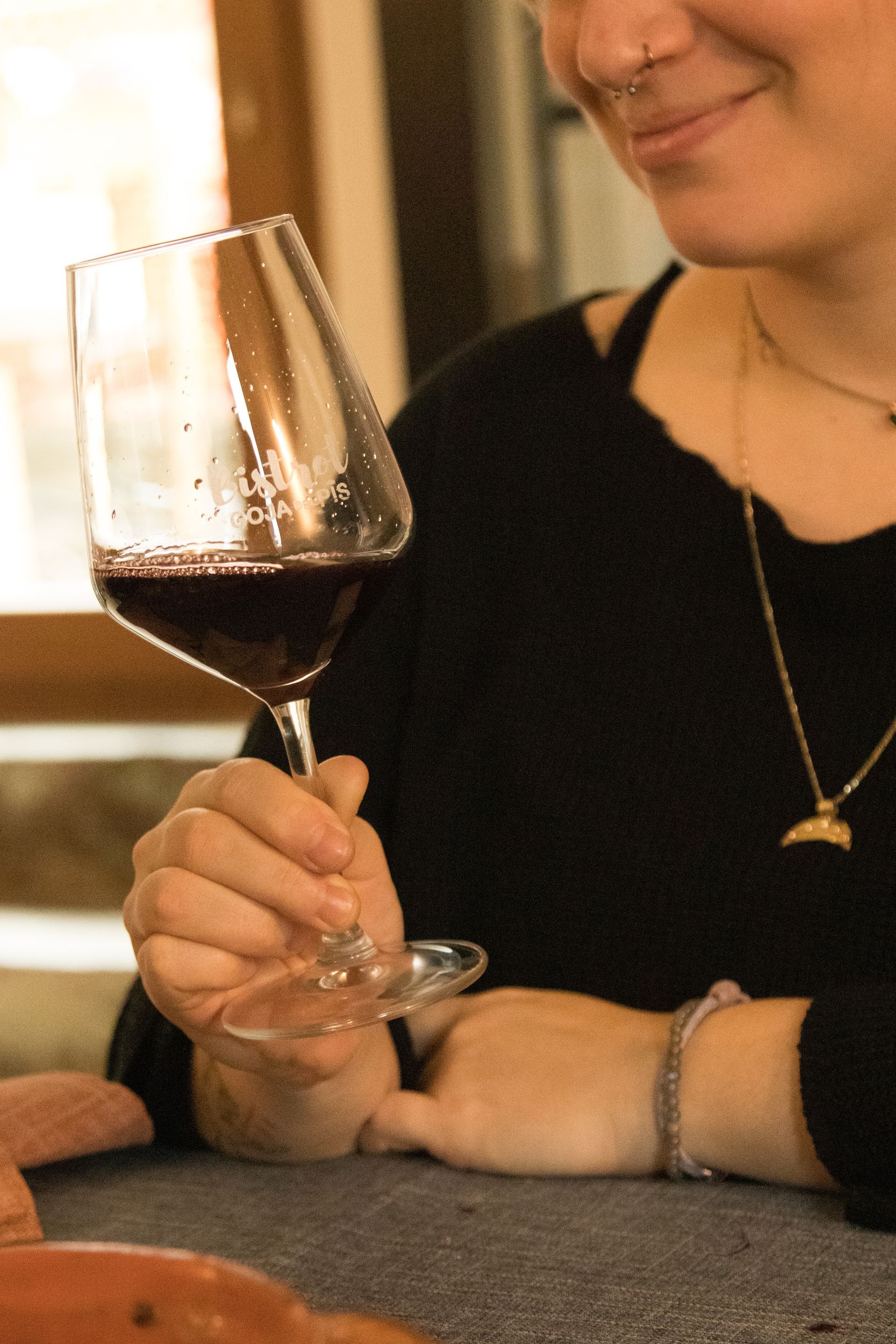 Una persona seduta a un tavolo tiene in mano un bicchiere di vino rosso e sorride guardandolo.
