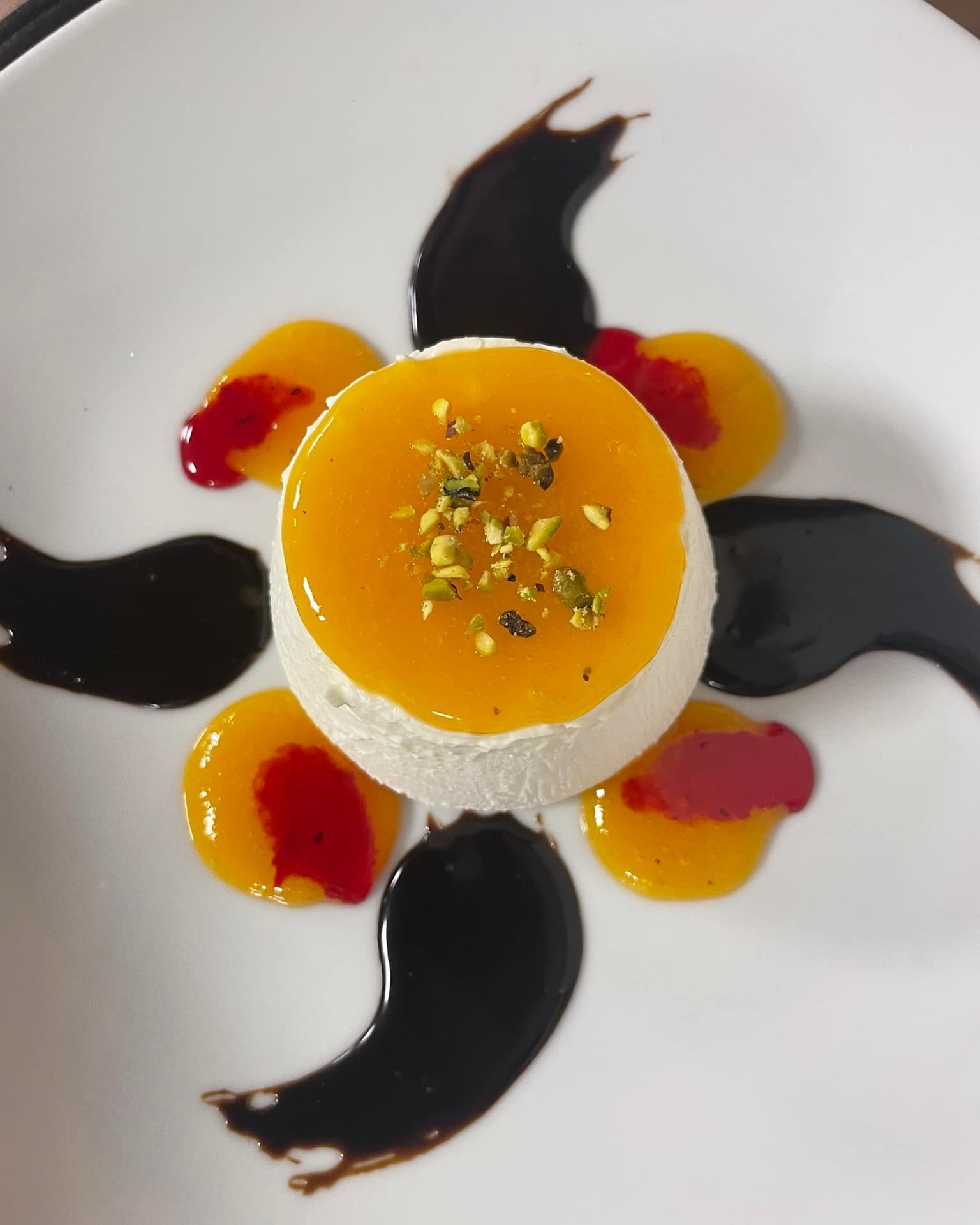 Dessert impiattato con crema bianca, ripieno di gelatina all'arancia, vortici di salsa nera e guarnizione in cima.