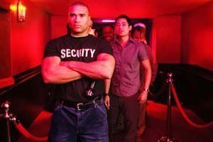 Security guard at club entrance, arms crossed, watching people in red-lit hallway.