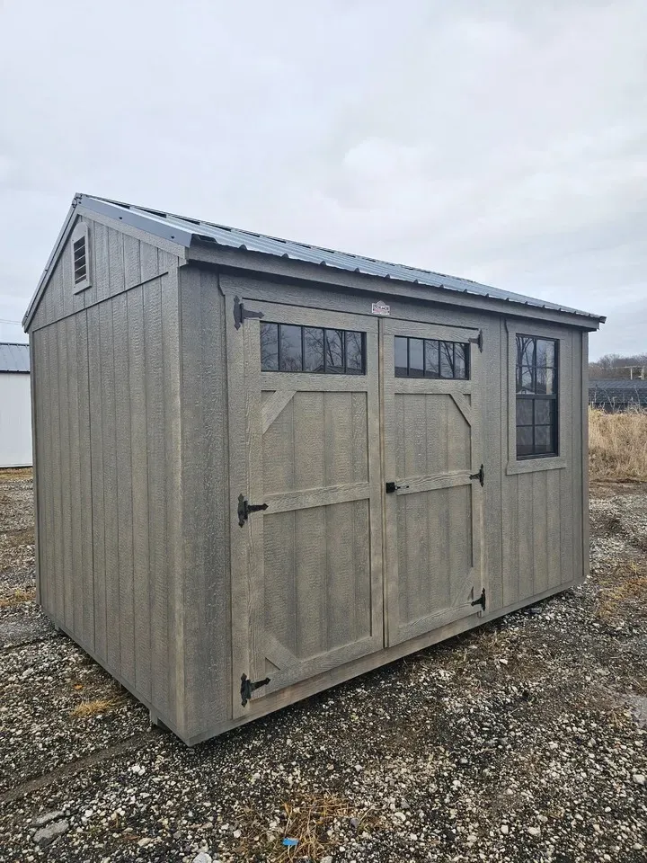 Weathered gray storage shed with double doors and small windows on a gravel lot under an overcast sky