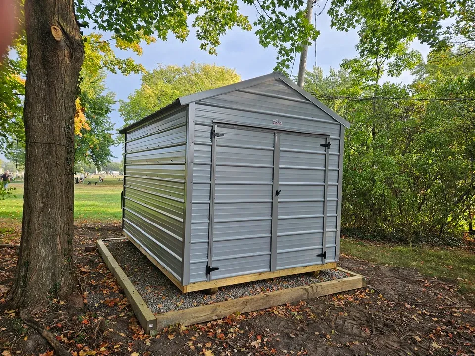 Gray storage shed on a concrete pad in a wooded yard, beside a tree and shrubs