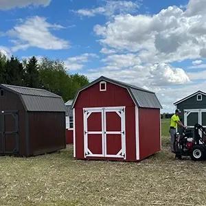Red and white shed in a grassy field, with another shed and a person on a lawn mower nearby under a cloudy sky