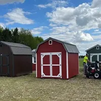Red and white shed in a grassy field, with another shed and a person on a lawn mower nearby under a cloudy sky
