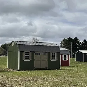 Green shed-style building with double doors in a grassy field, with a small red outbuilding behind it under cloudy skies