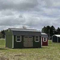 Green shed-style building with double doors in a grassy field, with a small red outbuilding behind it under cloudy skies
