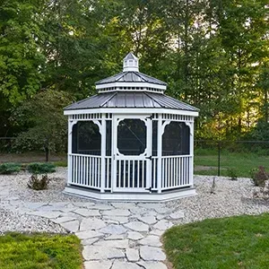 White octagonal gazebo with screened panels in a wooded backyard, surrounded by stone and gravel landscaping