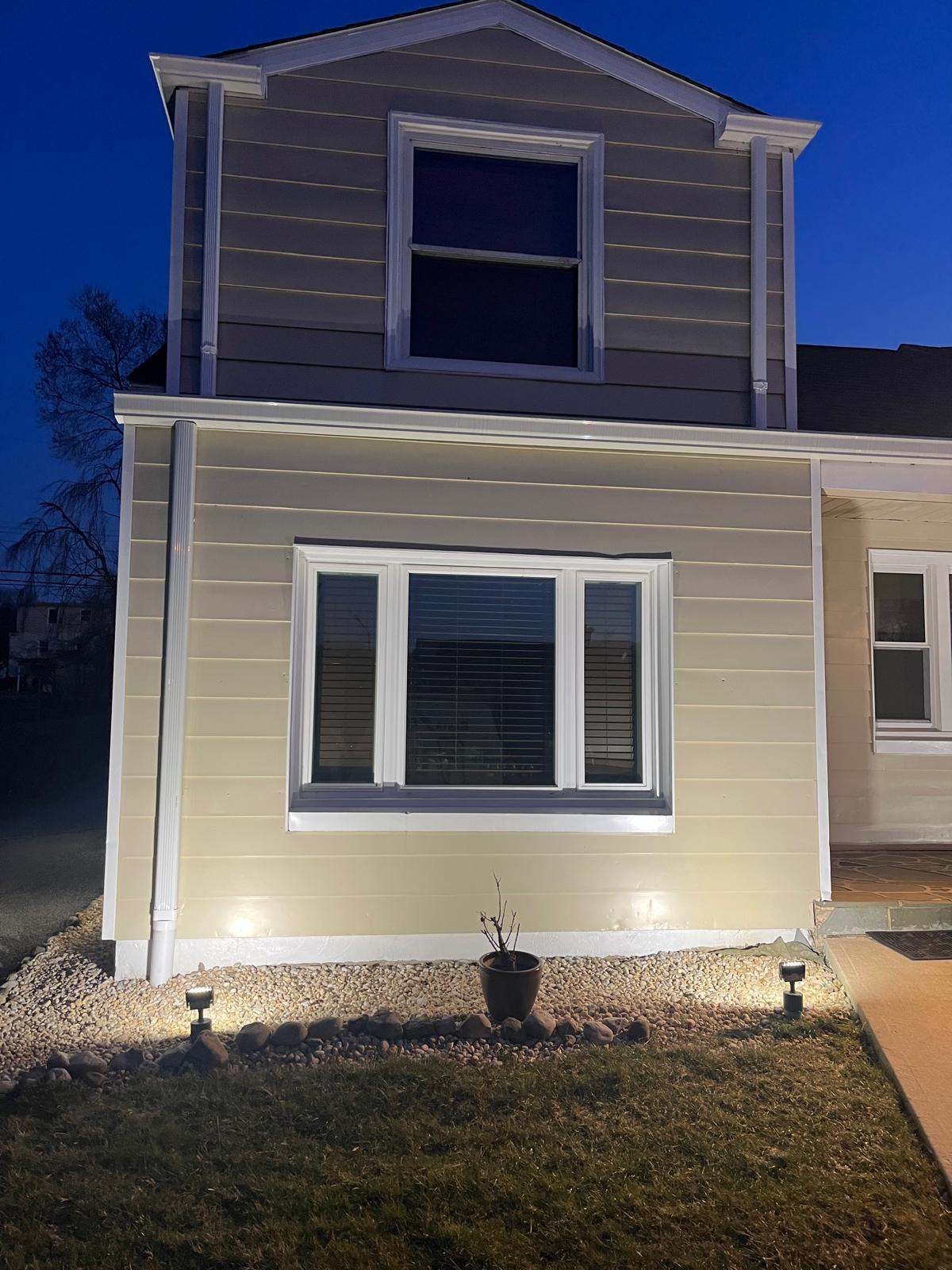 Lit beige two-story house at night with exterior lights illuminating the front windows and siding