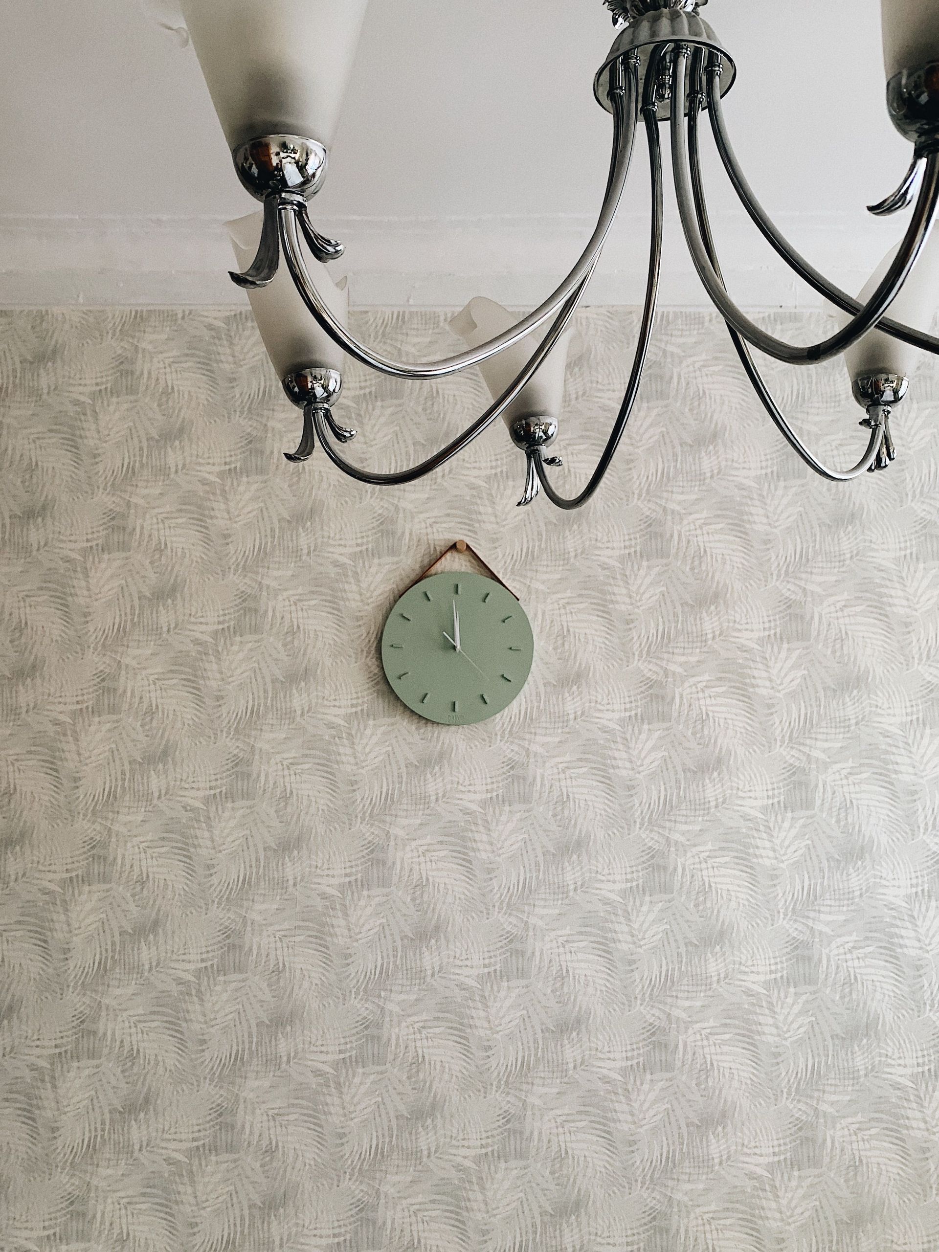 Green wall clock beneath a black chandelier on patterned wallpaper in a room.