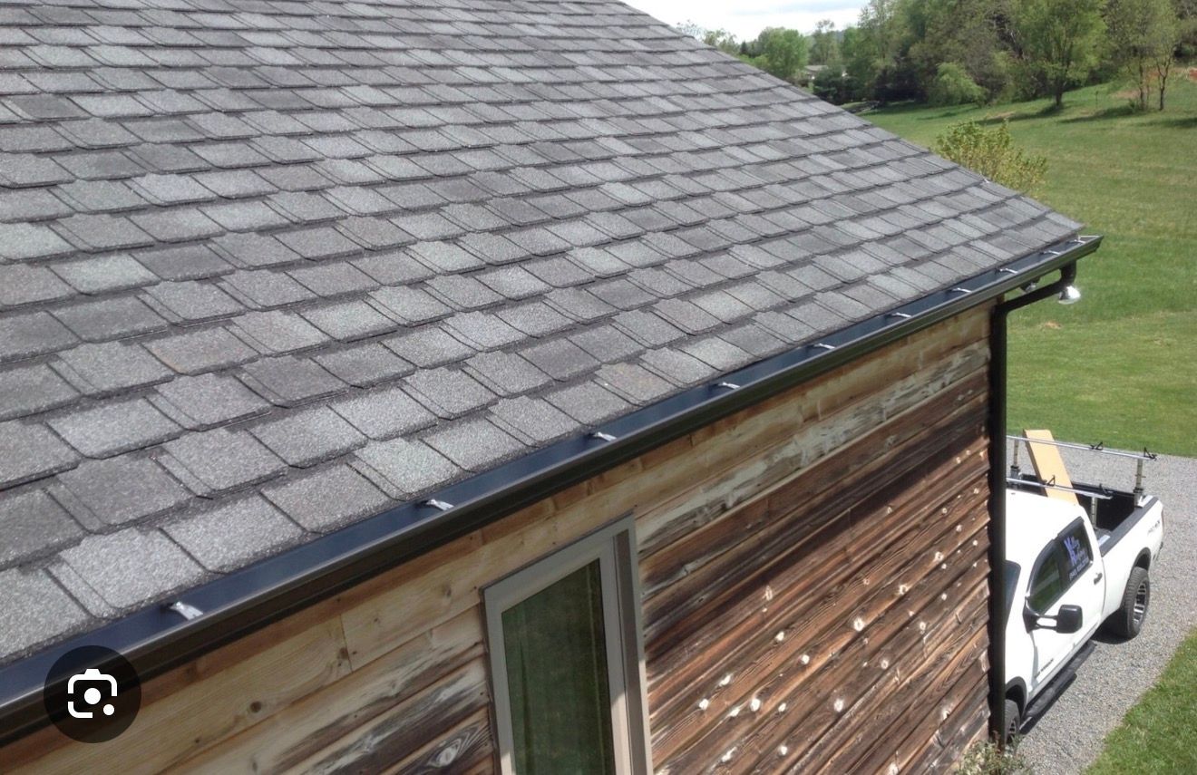 Roof edge with gray shingles above a wooden building and a white truck below on a grassy property.