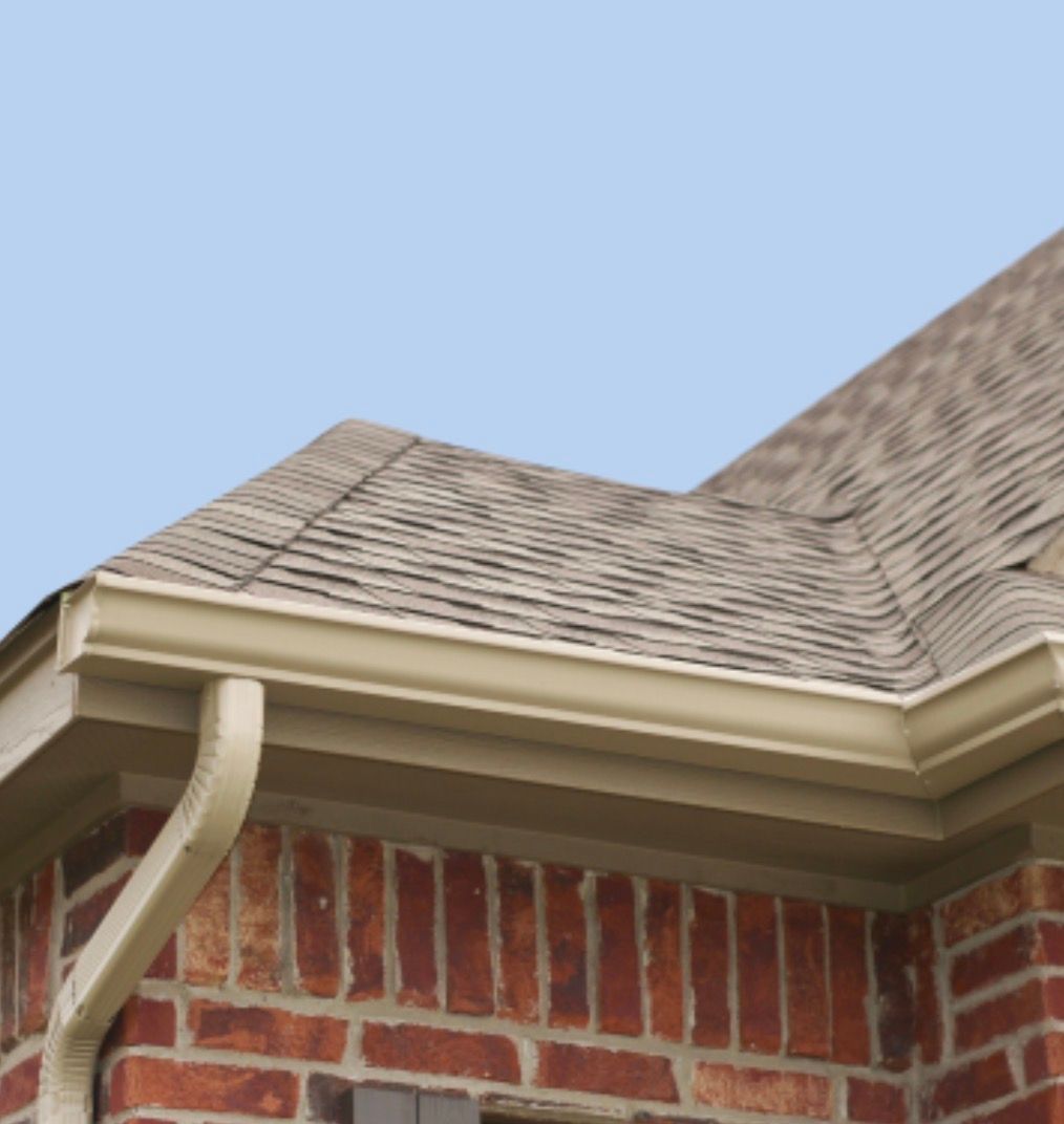 Close-up of a house roof with tan shingles, white trim, and red brick wall against a blue sky