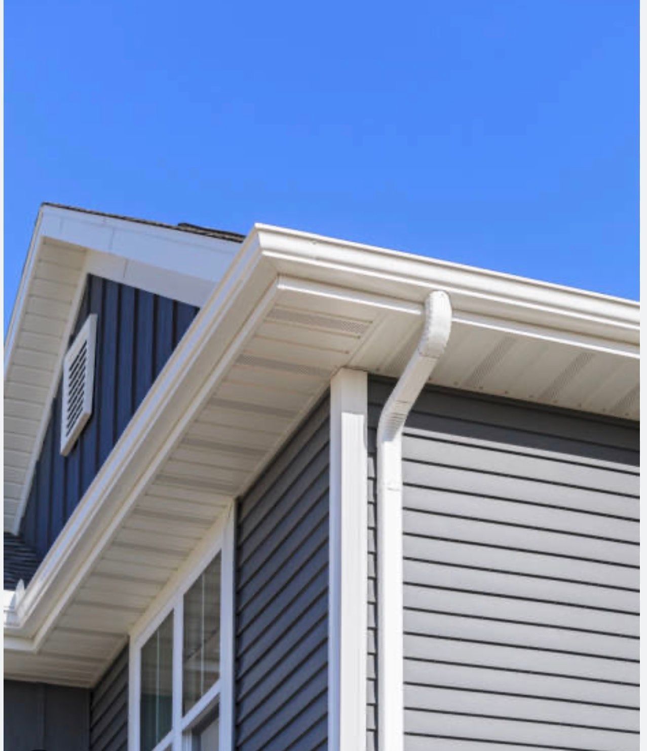 Close-up of a gray house corner with white trim, windows, gutters, and blue sky above.