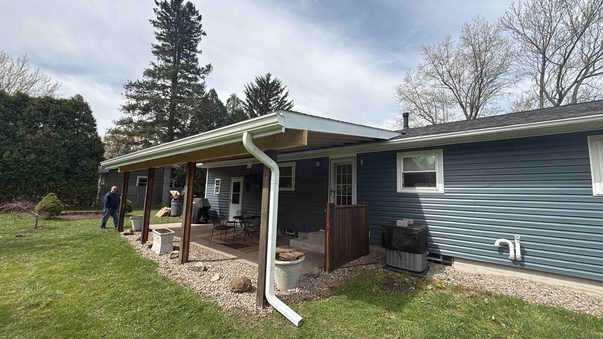 House with gray siding and covered porch on a grassy yard, with two people standing near the left side.
