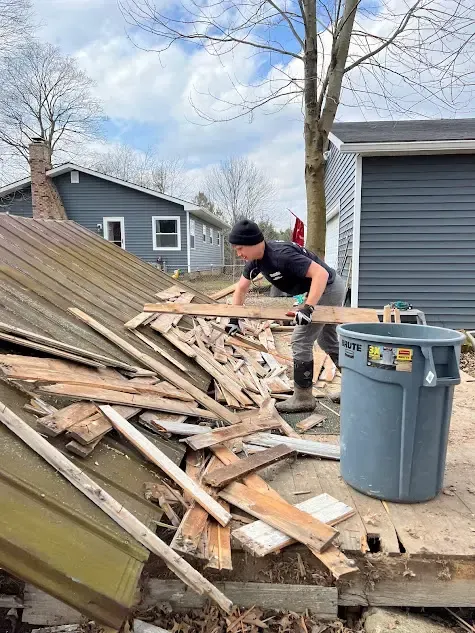 A man is standing on top of a pile of wood next to a trash can.