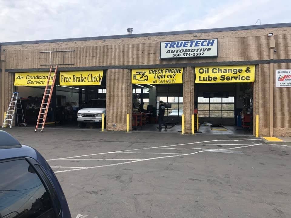 Auto repair shop storefront with yellow service signs, ladders, and parked cars in front | TrueTech Automotive