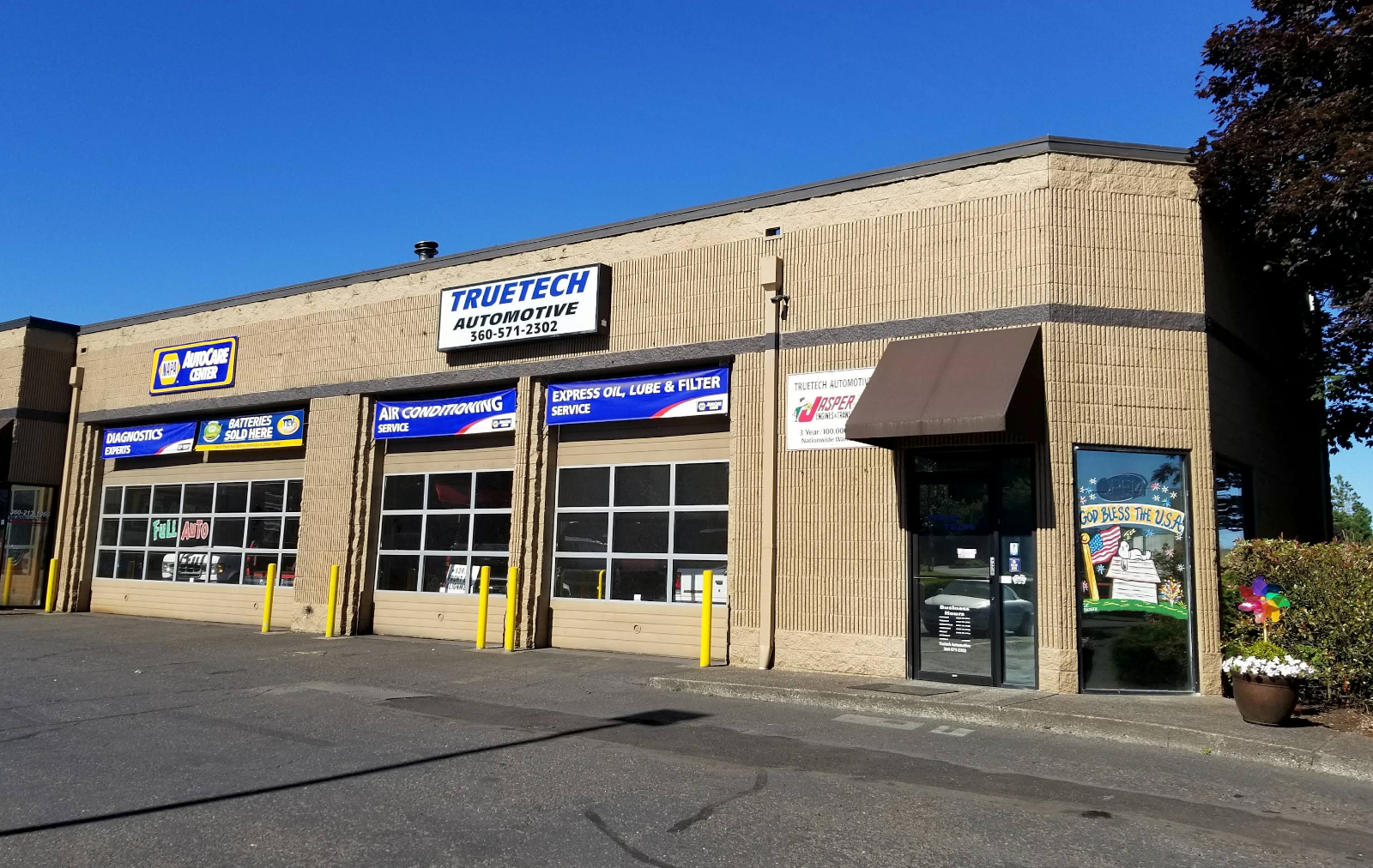 Small auto repair garage with three bay doors and signs on a sunny street corner | TrueTech Automotive