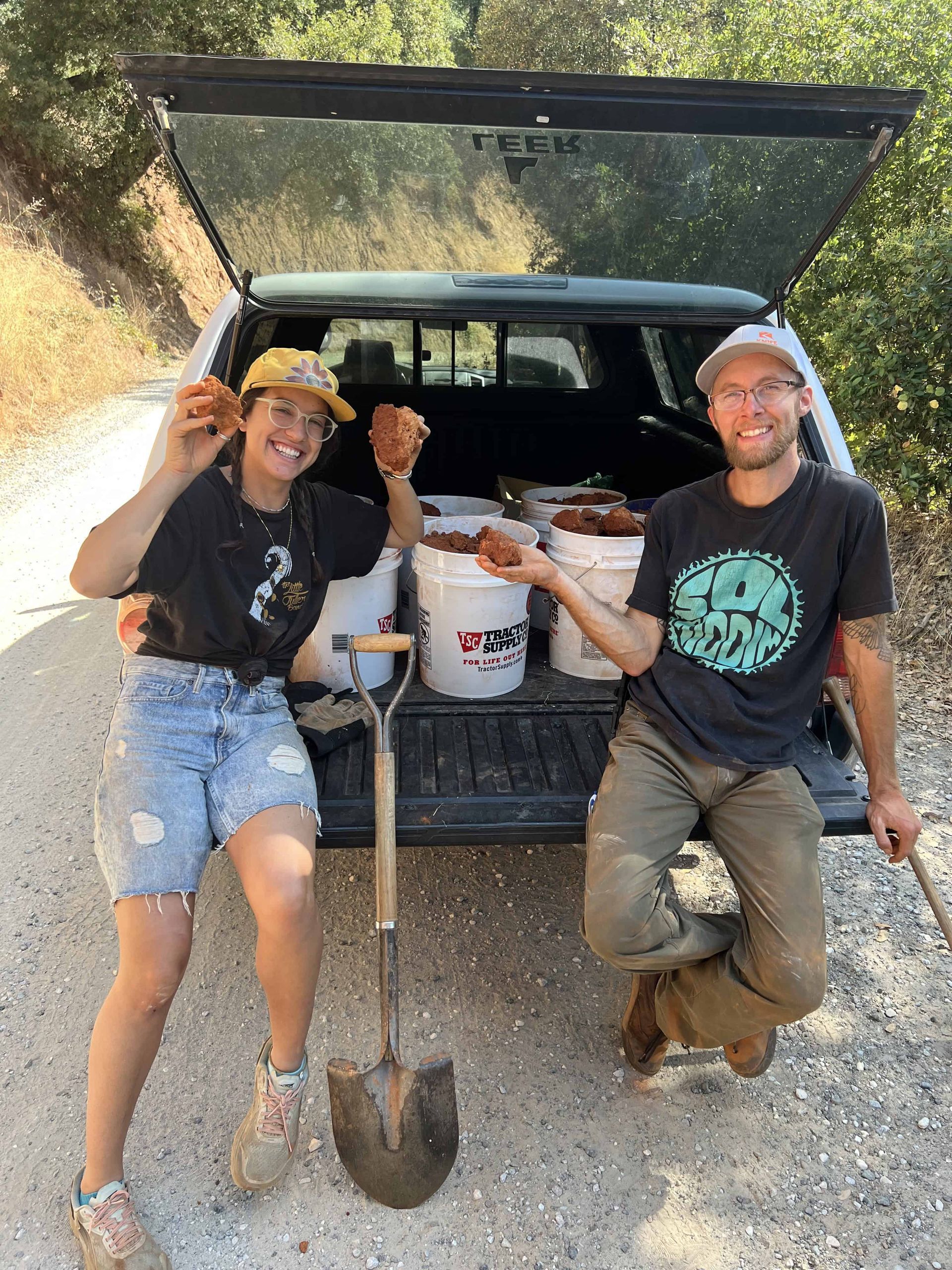 Smiling pottery students sitting on a truck tailgate, holding raw red earth they just dug up
