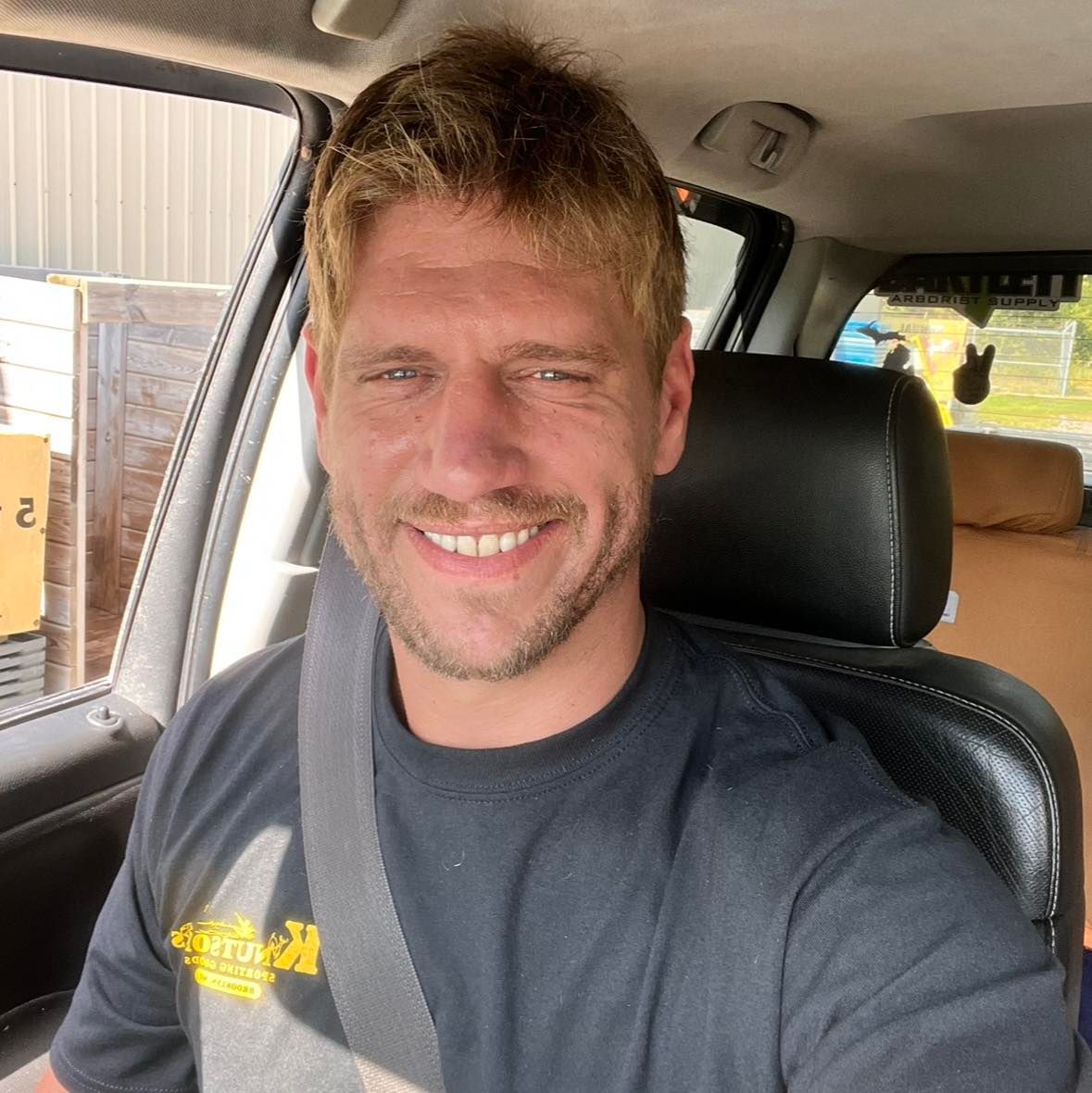 A man wearing a hard rock t-shirt is smiling while sitting in a car