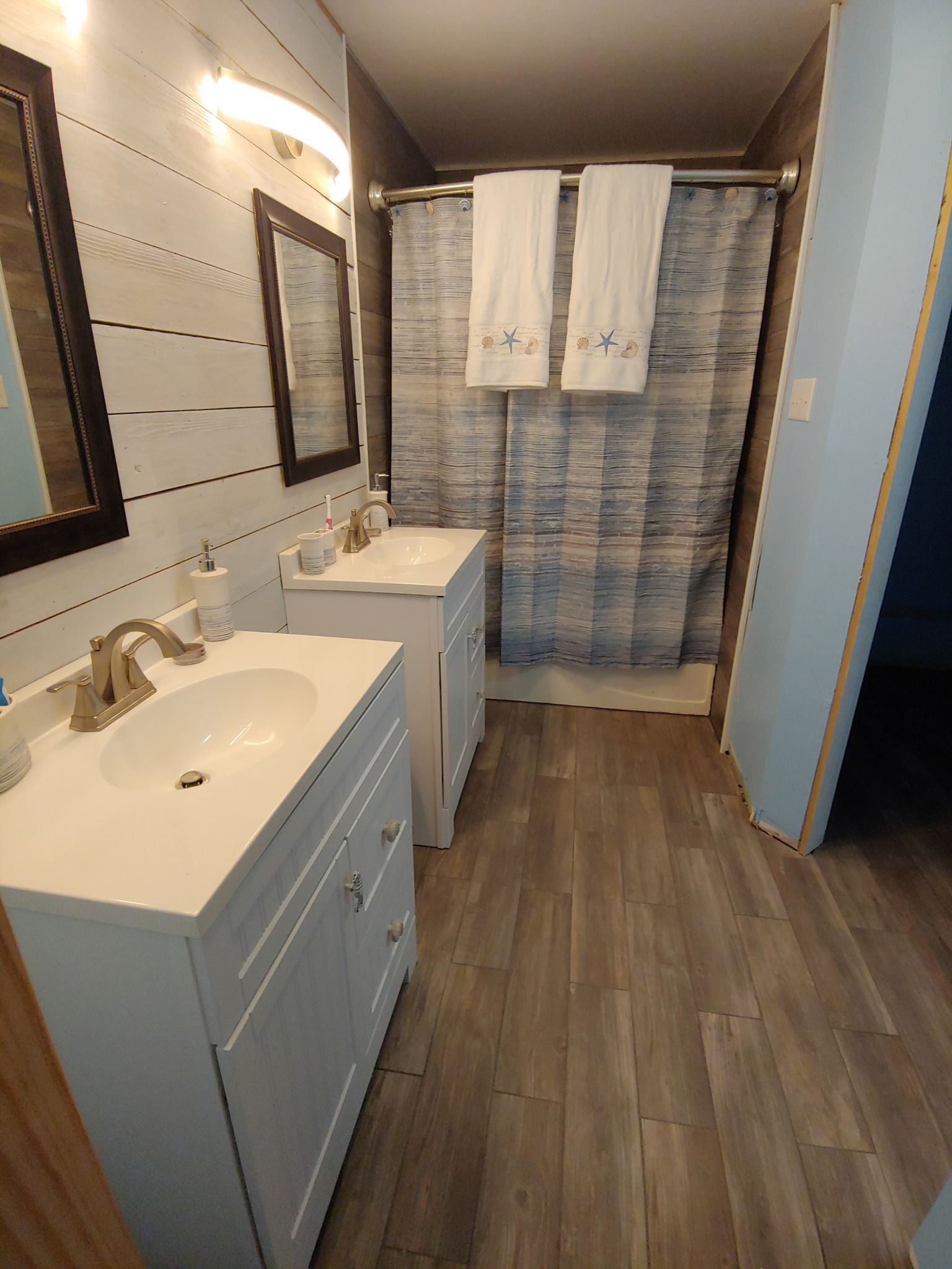 Bathroom with two white vanities, wood-look floor, shiplap wall, blue shower curtain, and dark-framed mirrors.