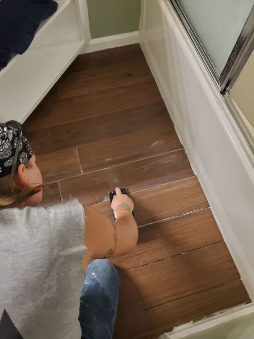 Person applying grout to wood-look tile floor in a bathroom corner.