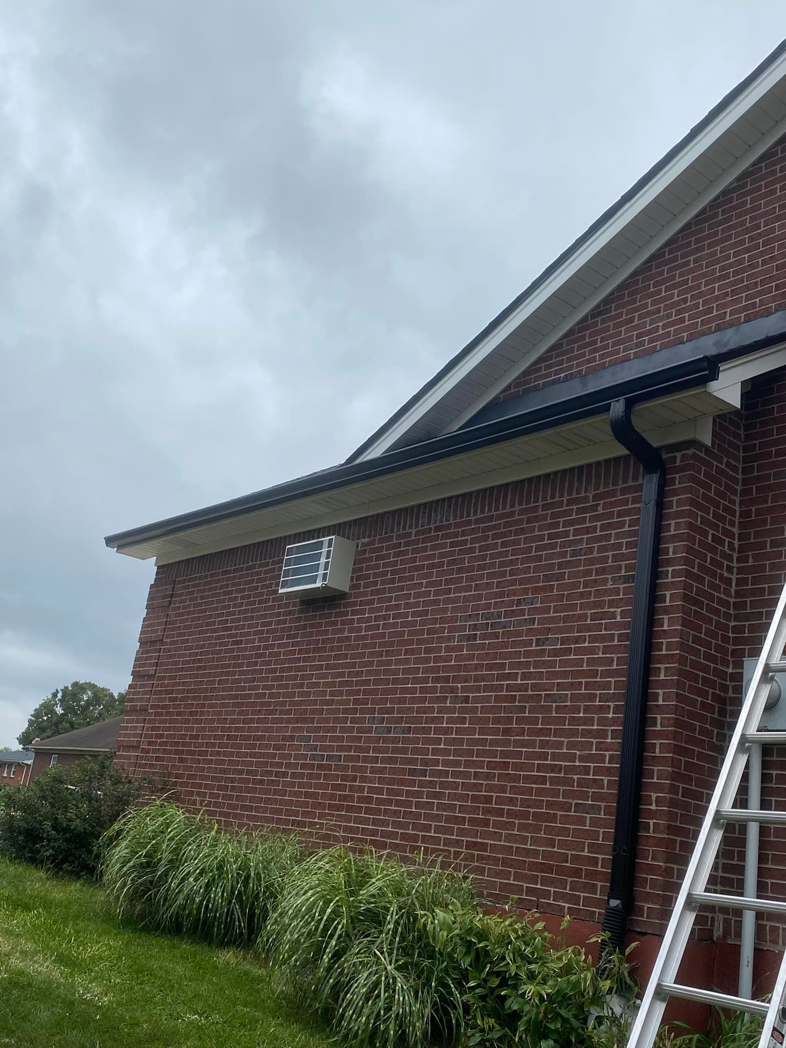 A side view of a red brick house exterior with a small vent near the roofline and a black gutter downspout beside a ladder.