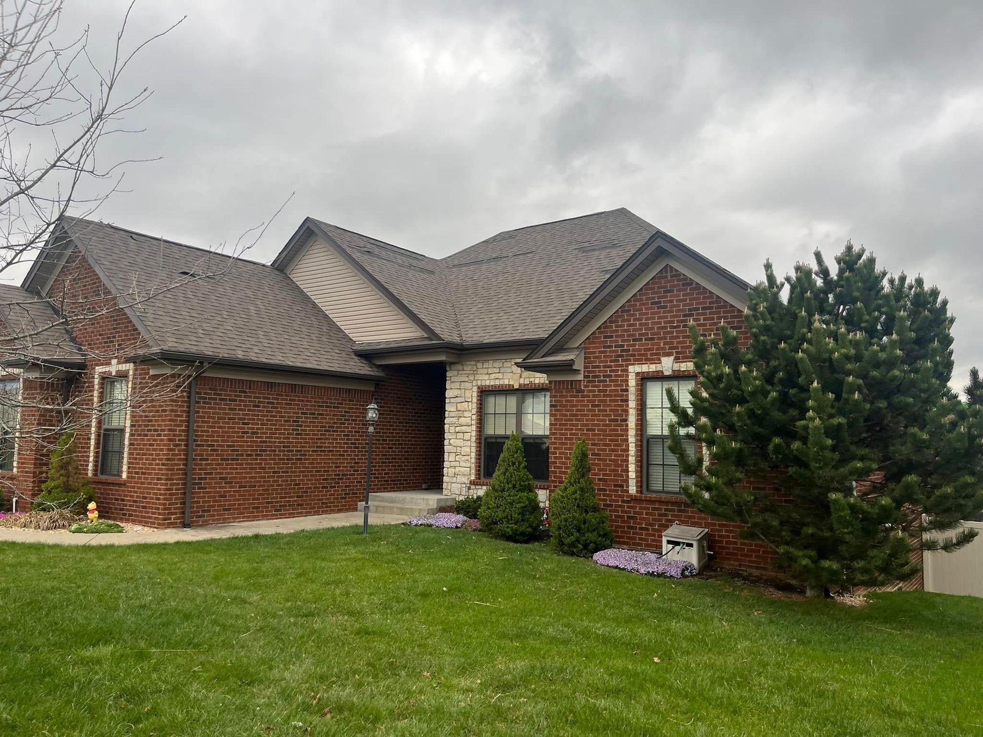 A one-story brick house with a stone accent and a gray shingled roof, surrounded by a lawn under a cloudy sky.