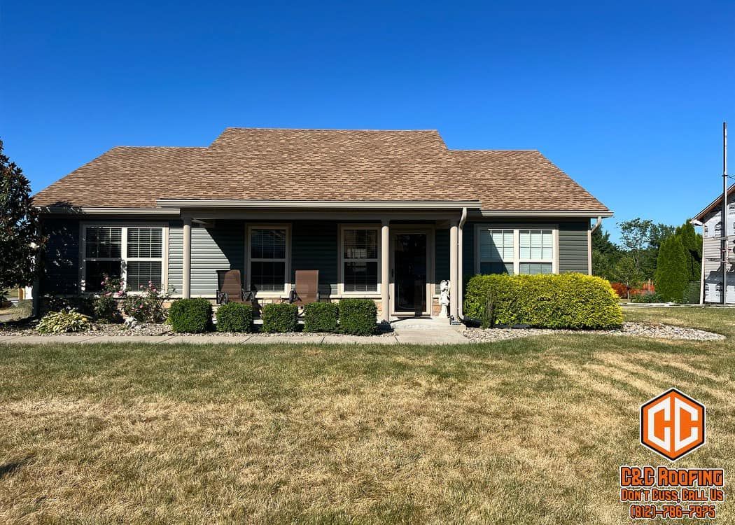 A one-story suburban home with a covered porch, tan siding, a brown shingled roof, and landscaped foundation shrubs.