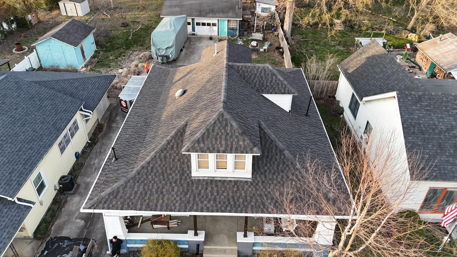 An aerial, high-angle view of a gray-roofed residential house, surrounded by neighboring homes and trees on a sunny day.