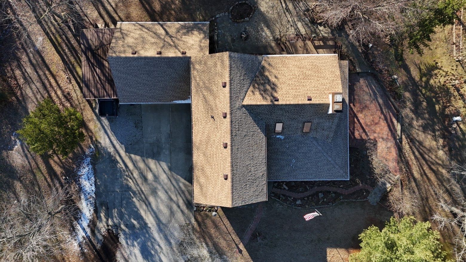 An aerial view of a suburban house with a brown shingled roof, surrounded by trees and a paved driveway.
