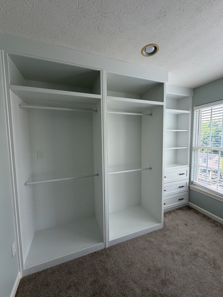 White built-in closet with open shelves and hanging rods in a carpeted bedroom, beside a window and drawer unit
