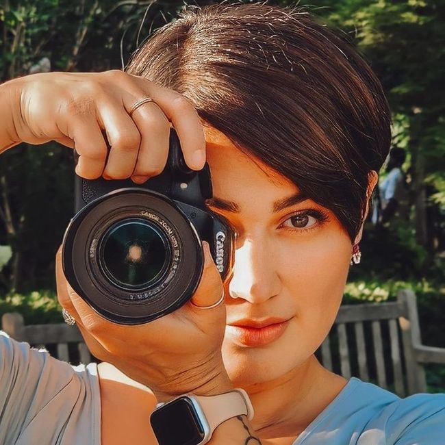Woman holding a Canon camera, outdoors. She is looking through the lens, with green foliage in the background.