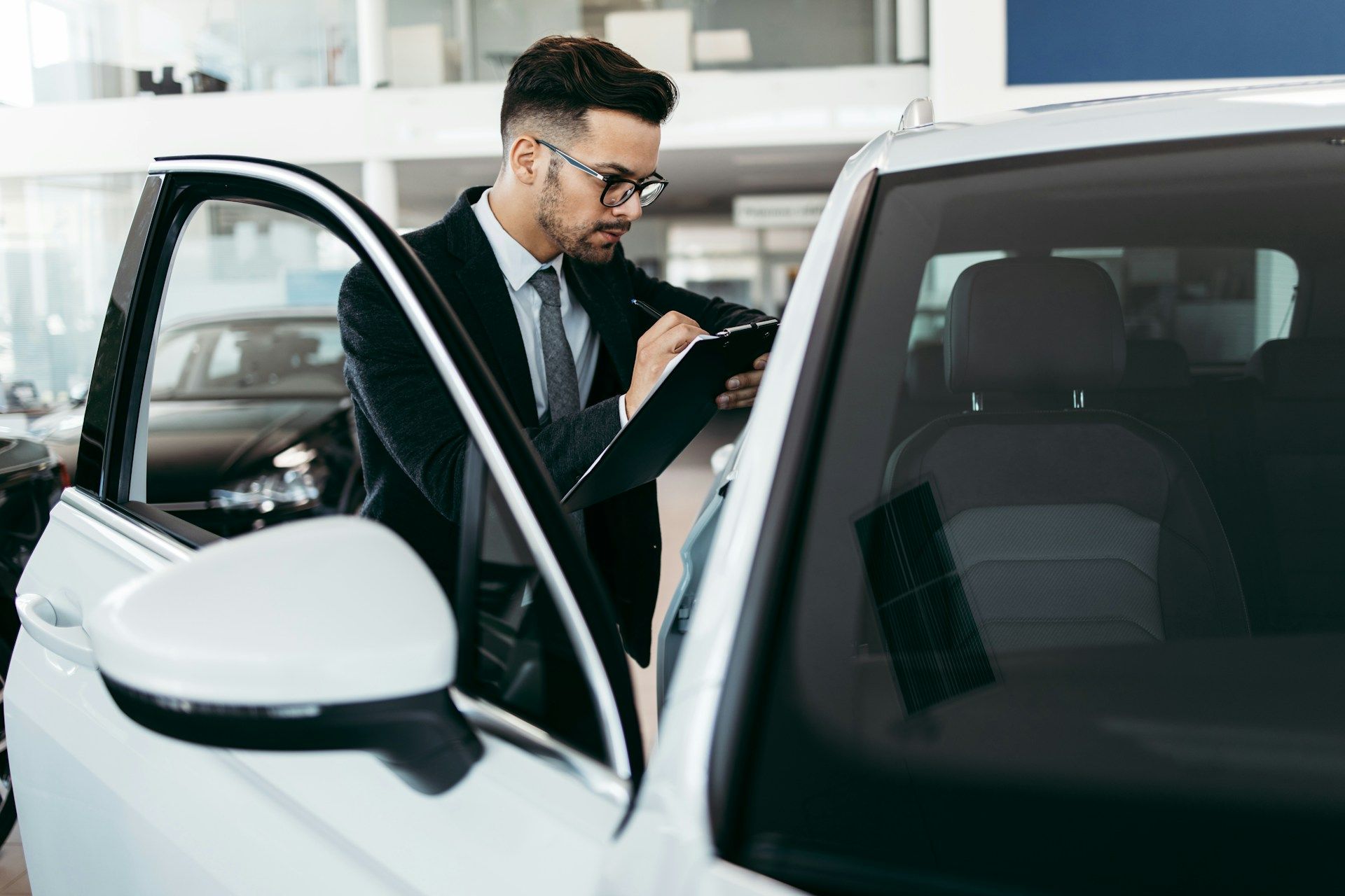 Car salesperson inspecting a white car in a dealership, taking notes on a clipboard.