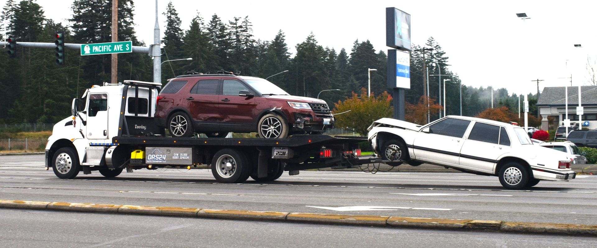 Tow truck carrying a maroon SUV and a white car on a road. Green trees and a sign are in the background.