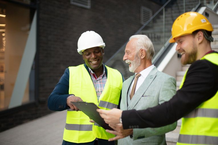 Three people, including two in hard hats and vests, looking at a clipboard outdoors.