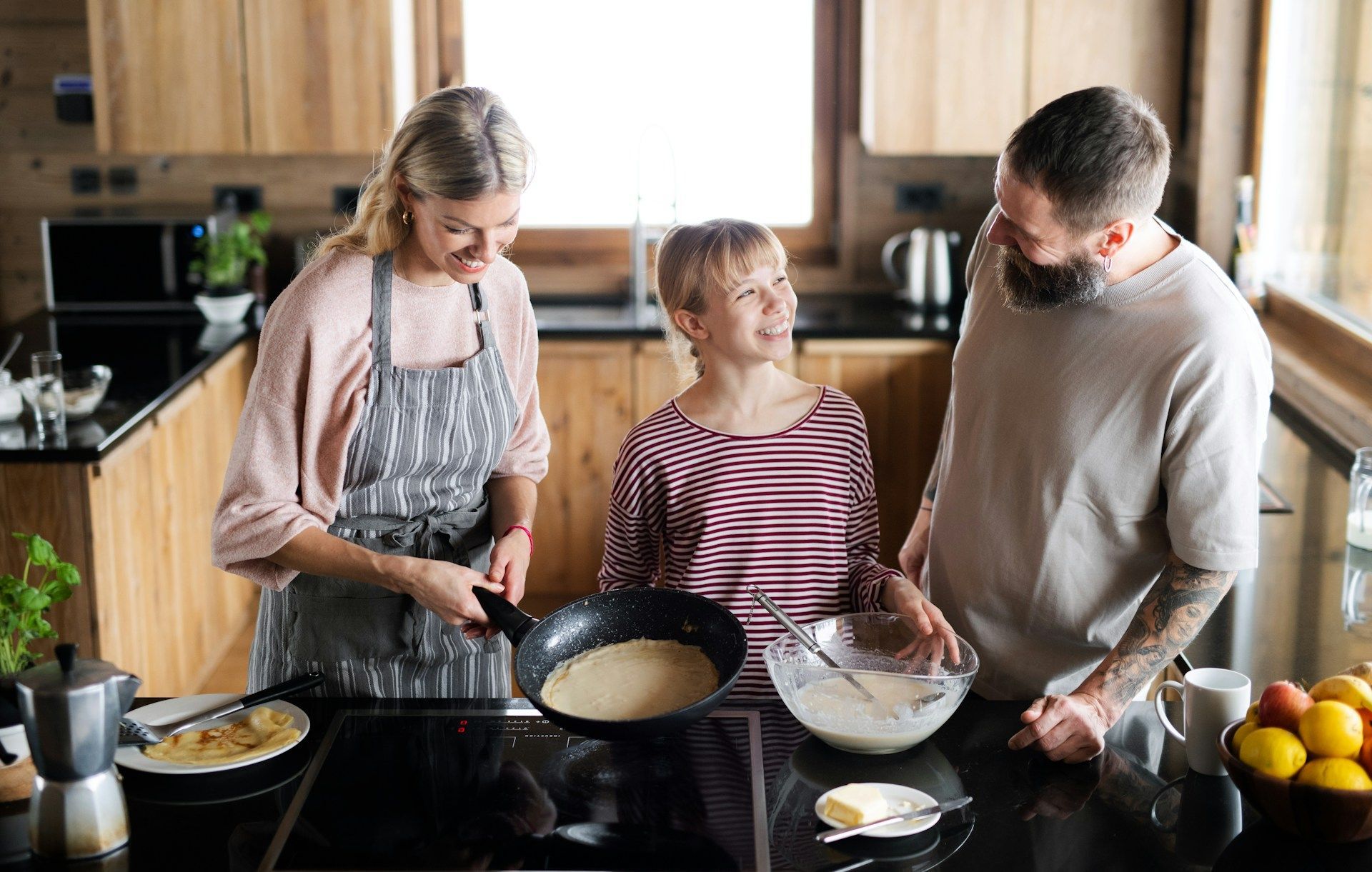 Family cooking pancakes in a kitchen; mother with apron flips a pancake, smiling at child who looks back at her, father watches.