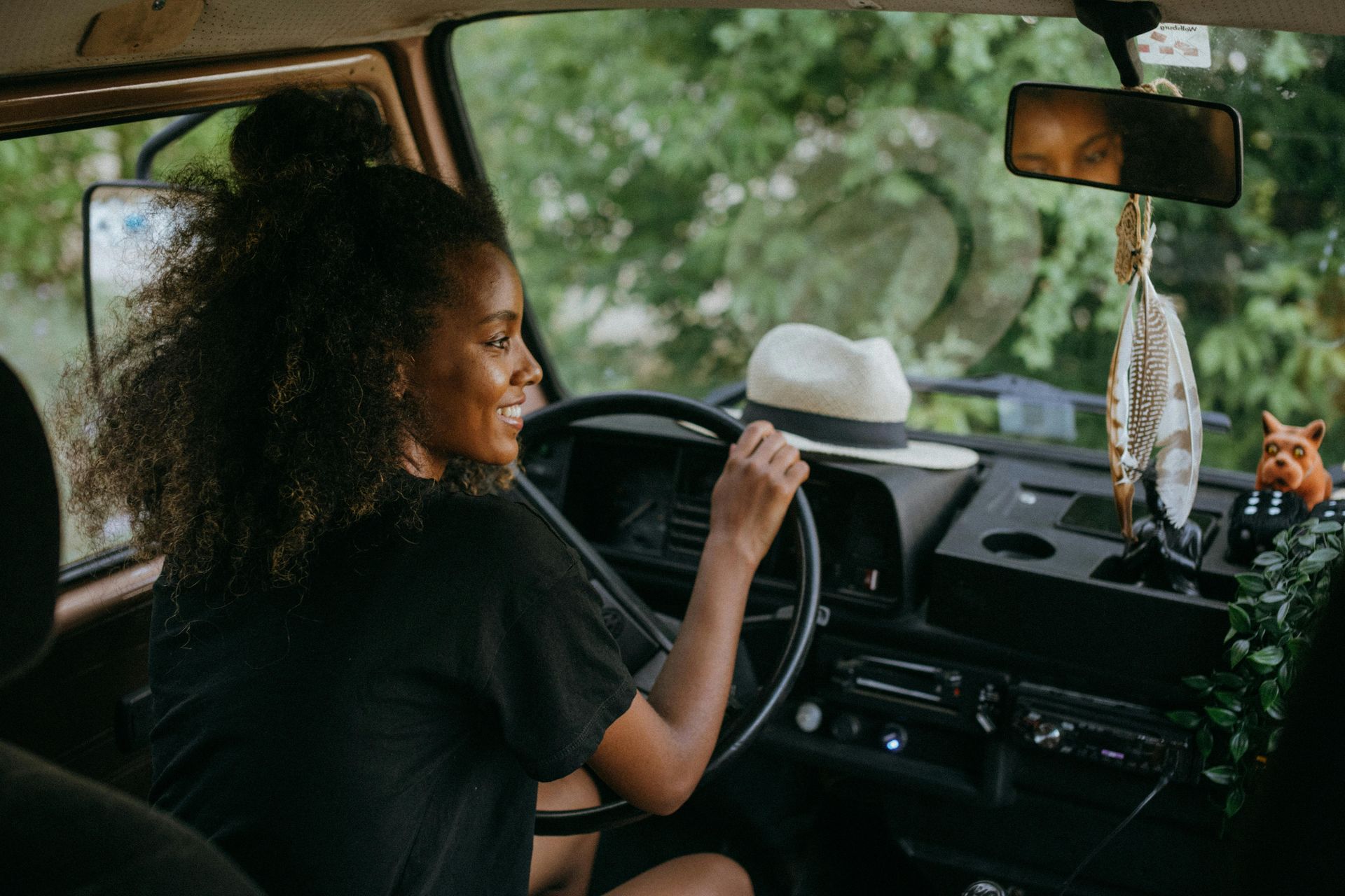 Woman smiles while driving a vintage van, surrounded by nature. A hat sits on the dashboard.