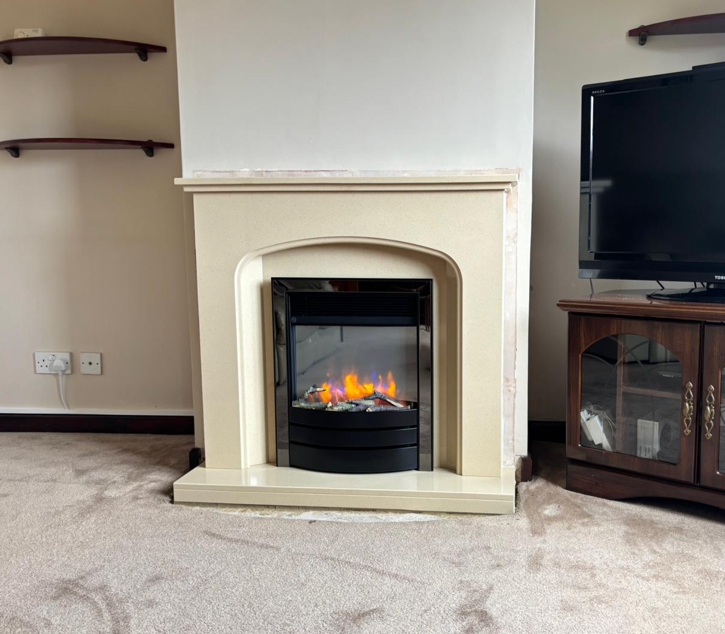 Living room fireplace with a lit electric flame, beige mantel, TV stand, and carpeted floor