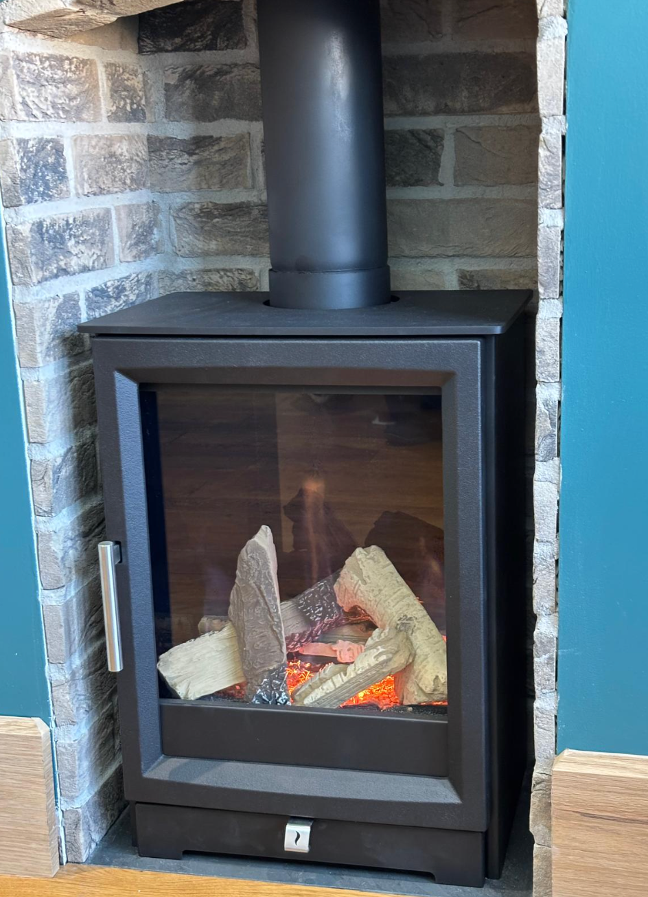 Black wood-burning stove with glass door and lit faux logs in a stone fireplace alcove