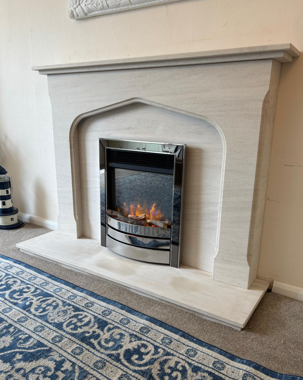 White fireplace mantel with a lit electric fire in a beige room, beside a blue patterned rug.